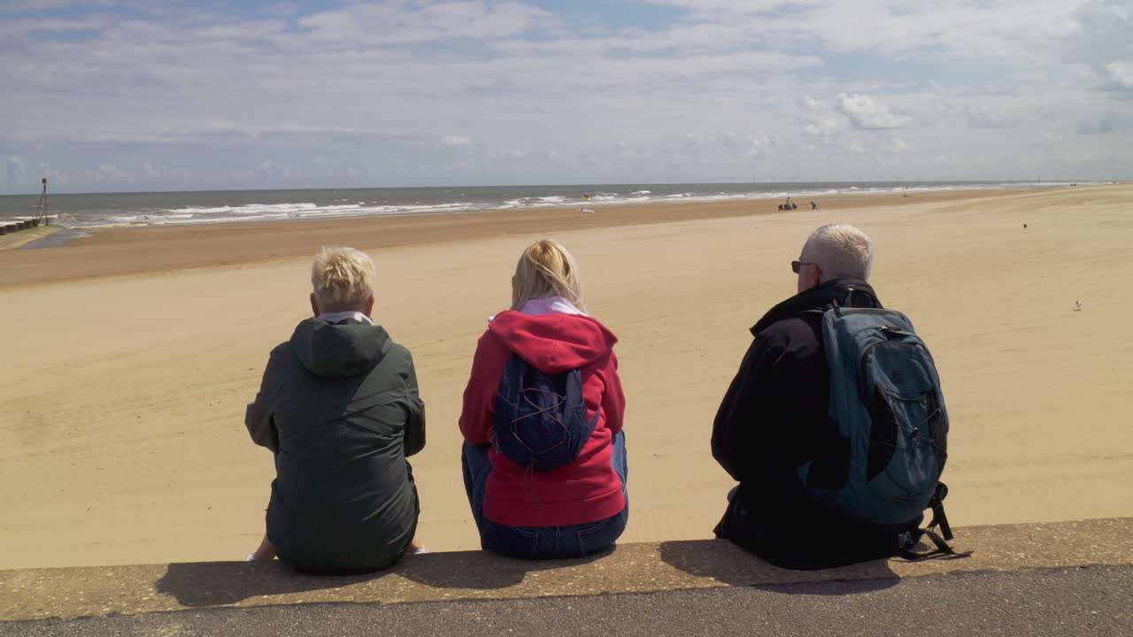 Three friends sit on a wall, taking in the beautiful view of the sea and sandy beach under a bright sky