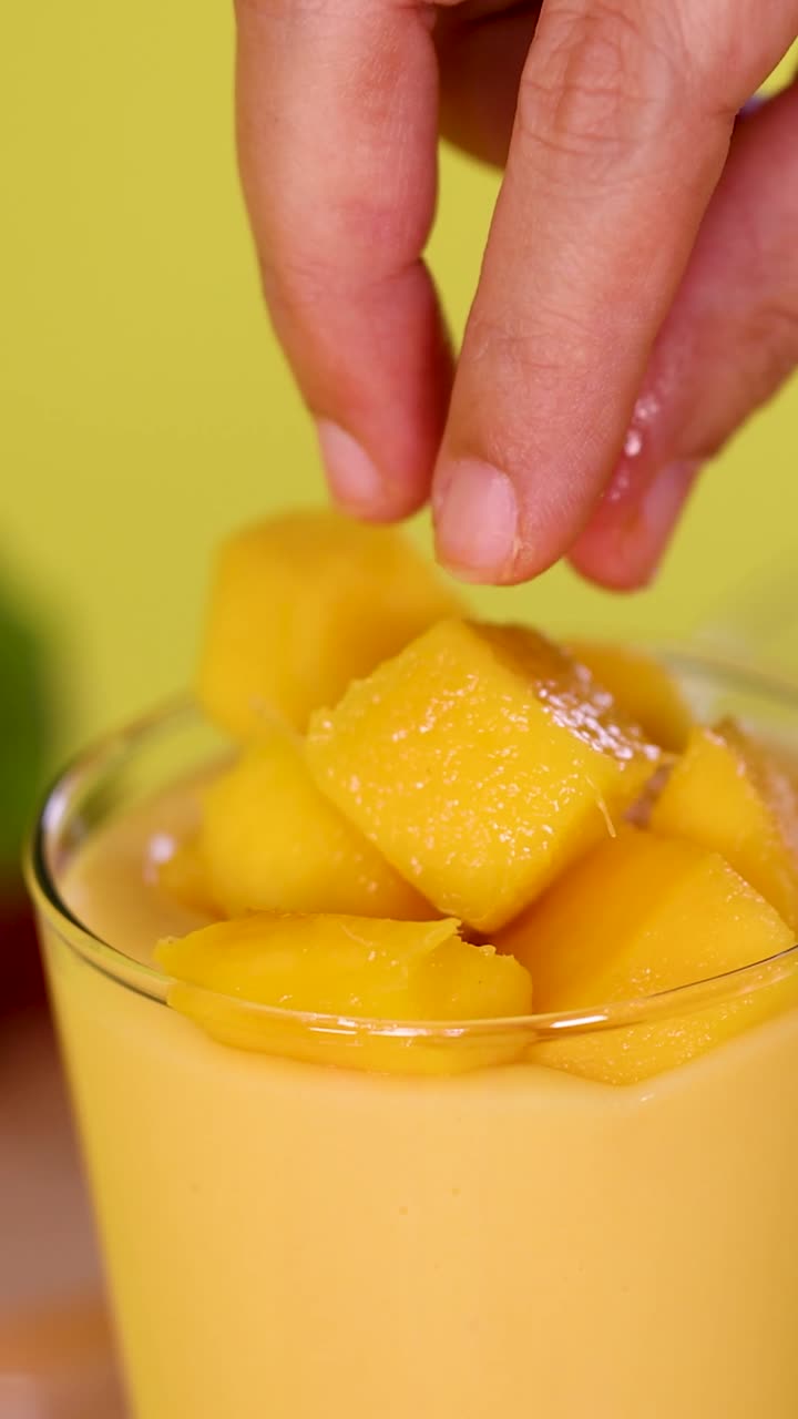 Una mano lascia cadere cubetti di mango fresco in un bicchiere giallo per frullati su uno sfondo luminoso