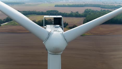 Nahaufnahme-Einer-Windturbine-Mit-Einer-Drohne,-Die-über-Die-Propeller-Aufsteigt-Und-Nach-Unten-Kippt,-Mit-Ackerfeldern-Im-Hintergrund