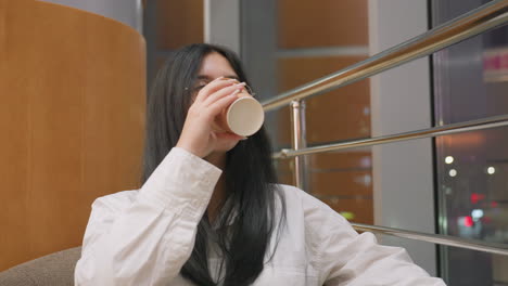 young woman with long dark hair and glasses sips coffee from paper cup while gazing outside through window with soft indoor lighting and warm evening city bokeh glowing in background