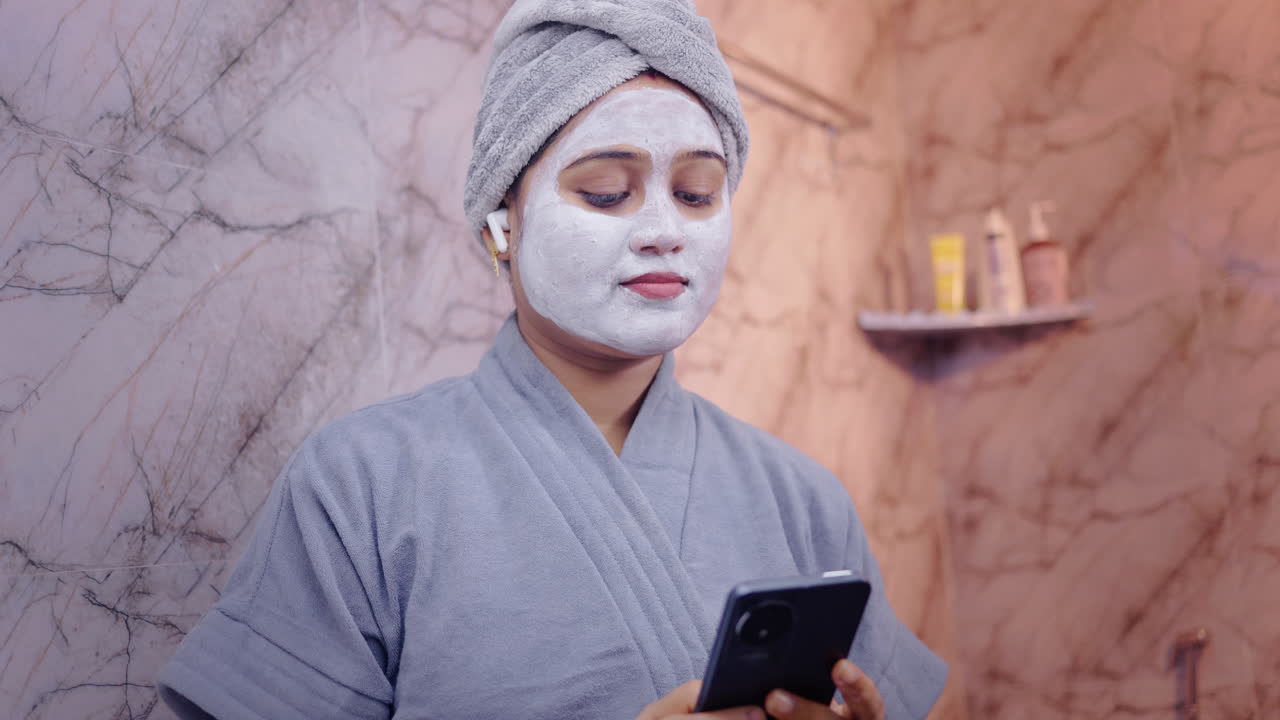 Donna che applica una maschera facciale e usa gli auricolari per ascoltare musica sullo smartphone, in piedi nel bagno moderno.