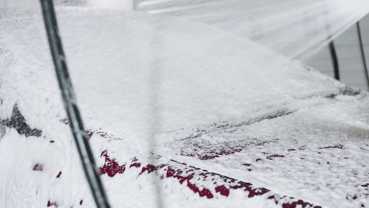 Close up of car washing process showing soapy foam and high pressure water spraying across windshield surface covering red vehicle with white bubbles