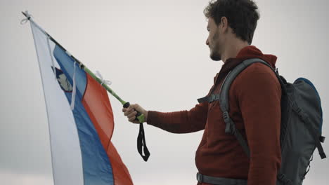 un joven excursionista con chaqueta roja y mochila gris sosteniendo un bastón de senderismo en el que colocó una bandera eslovena y ondea en el viento