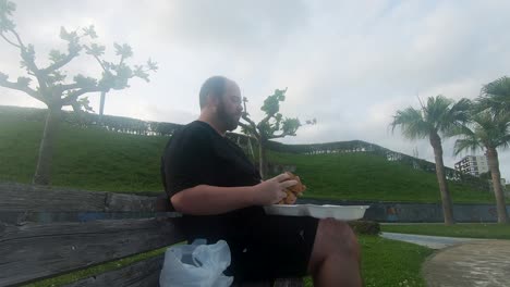 unfit man contributing to his poor health by eating a burger alone on a park bench