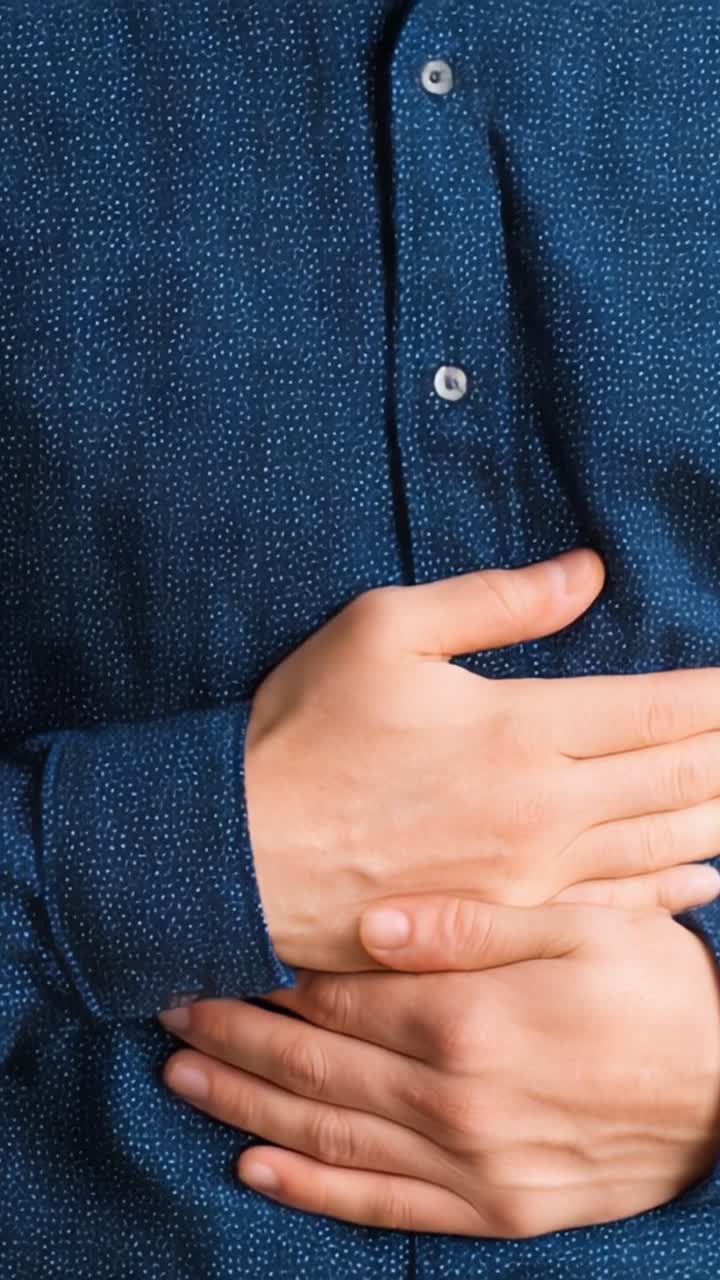 A close-up view of a person's hands clasped over their stomach, indicating discomfort or pain while wearing a stylish dark blue shirt with a dotted pattern