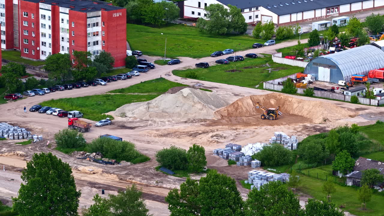 Open construction site with sand piles and equipment near residential buildings and greenery