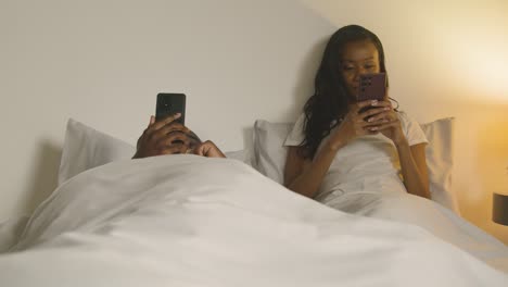 couple with relationship problems at home at night lying in bed both looking at mobile phones