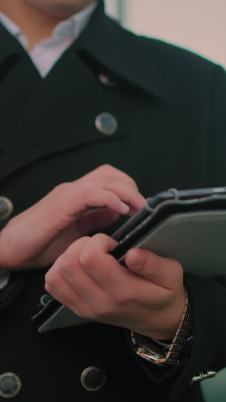 primer plano de una persona con abrigo negro operando una tableta cerca de un edificio de vidrio, reflejando coches estacionados y figuras borrosas en el vidrio, mostrando el entorno de la ciudad moderna