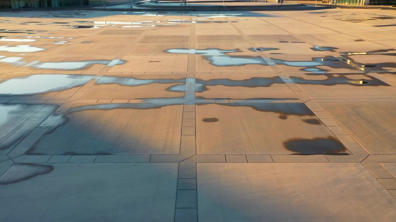 Vista angolata dall'alto di un'ampia copertura realizzata con pannelli rettangolari di cemento, con pozzanghere poco profonde, chiazze bagnate lucide, riflessi tenui e luce solare calda che creano una texture urbana geometrica e calma.