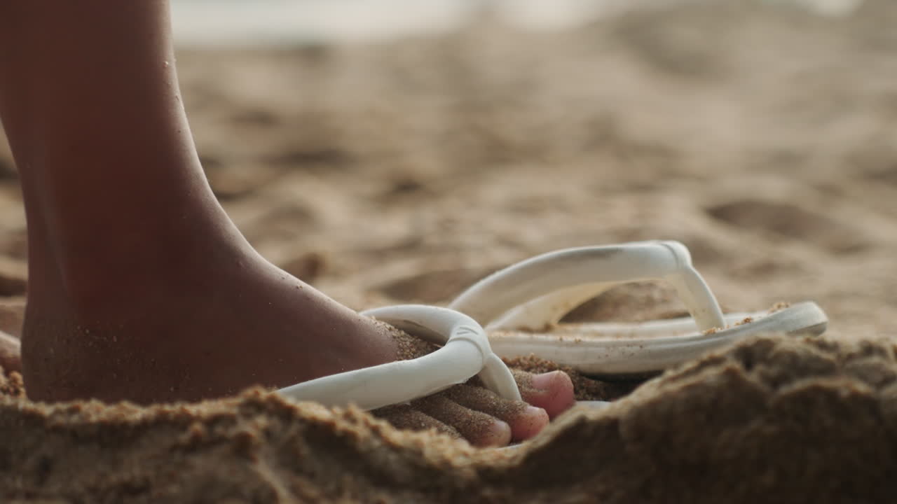 Bar fot lyfts från sandig sandal, snabb närbild på tår som släpper vit rem, sandkorn sprids när foten lyfts, lekfull rörelse och energisk rytm, barn eller tonåring som hoppar längs strandlinjen, varmt