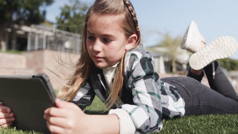 young girl using tablet outdoors, lying on grass, focused on screen, at school