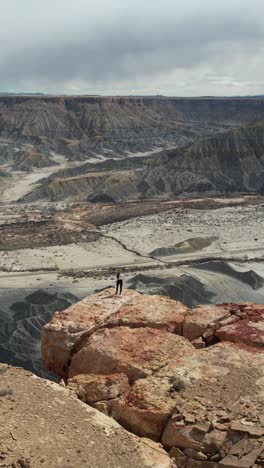 Vertical-Drone-Shot-of-Woman-Standing-on-The-Edge-of-Cliff-Above-Abyss-and-Desert-Landscape