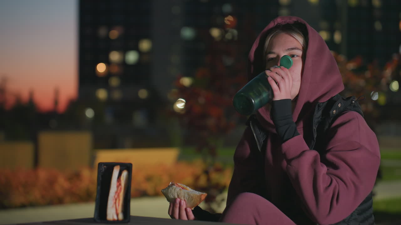 Young woman in hoodie sits outdoors at dusk under bright city lights eating sandwich while sipping water from bottle beside snack tray on wooden table in urban park breathing in crisp cool air