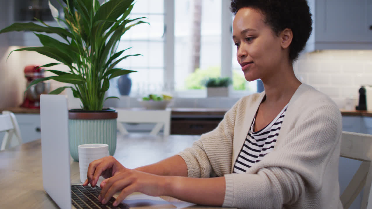 Mixed race woman using laptop and drinking coffee in kitchen