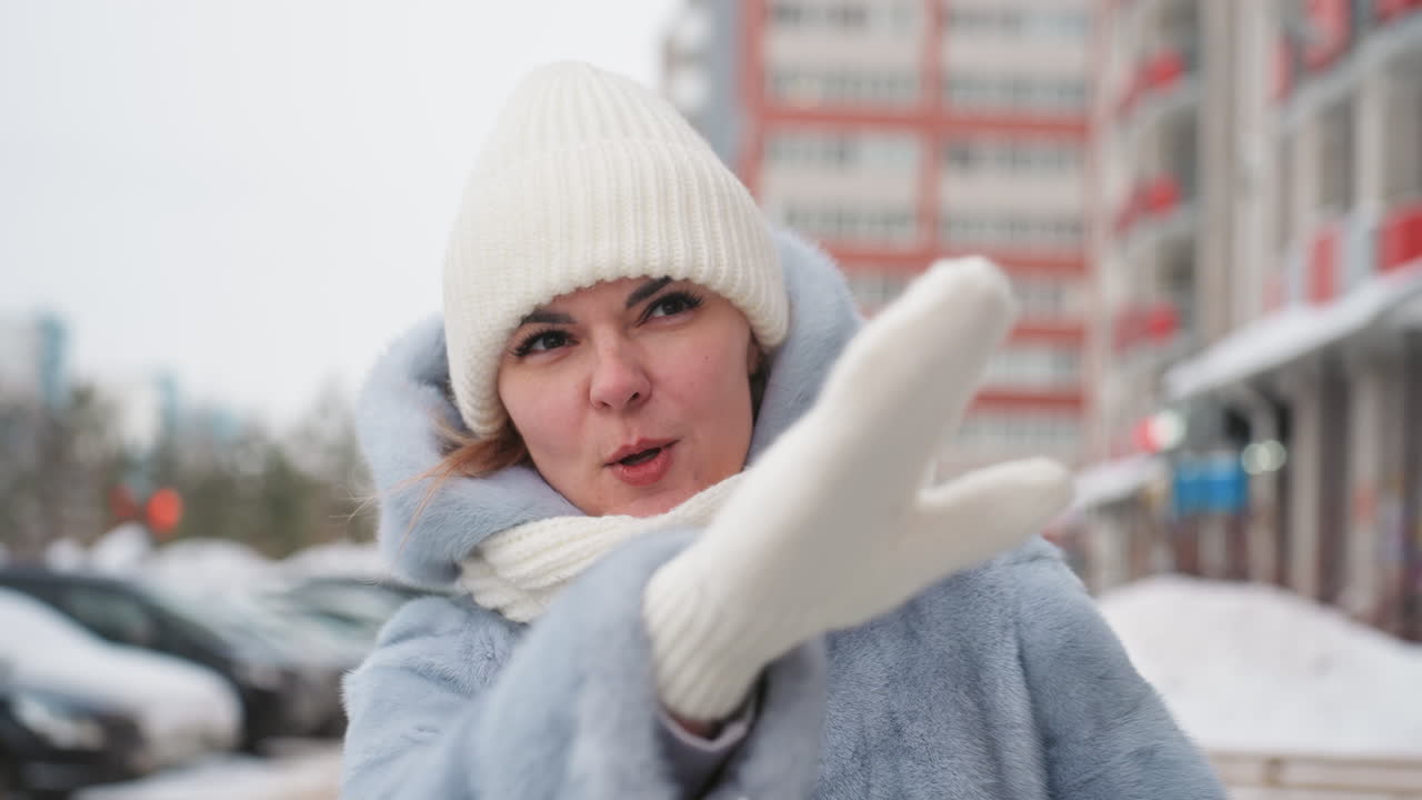 Caucasian mom calling son on snowy city sidewalk, wearing white knit hat, cream scarf and blue coat. Expressive hand gestures and concerned facial expression, visible breath in cold air. Urban