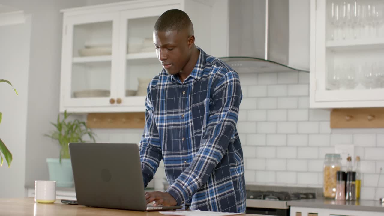 African american man using laptop and talking on smartphone in kitchen at home, slow motion