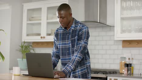 african american man using laptop and talking on smartphone in kitchen at home, slow motion