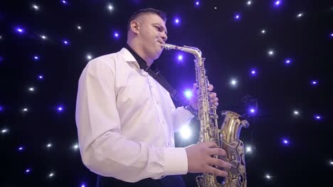 tilt up shot of biracial jazz musician in checkered suit and bowtie playing saxophone while sitting against black band member in dimly lit bar, rehearsing and checking sound before live performance