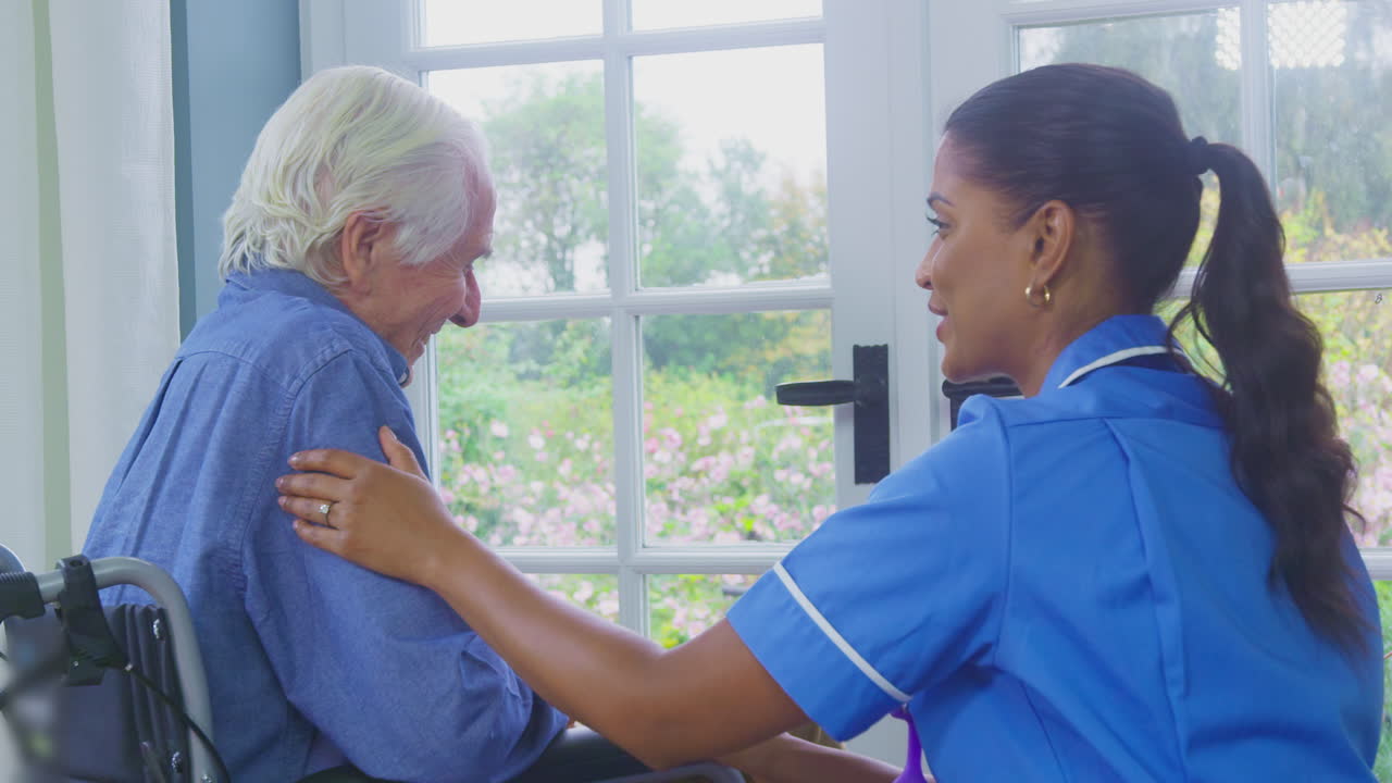 Female Care Worker In Uniform Talking With Senior Man Sitting In Wheelchair In Care Home Lounge