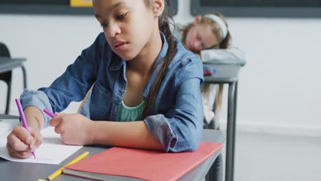 Video-of-bored-biracial-schoolgirl-sitting-at-desk-writing-in-class,-copy-space