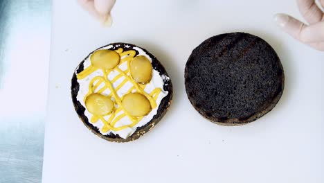 Amateur chef making hamburger. Cook puts pickles on black bun with sauce for preparing burger. Detailed process of cooking healthy burger. Top view