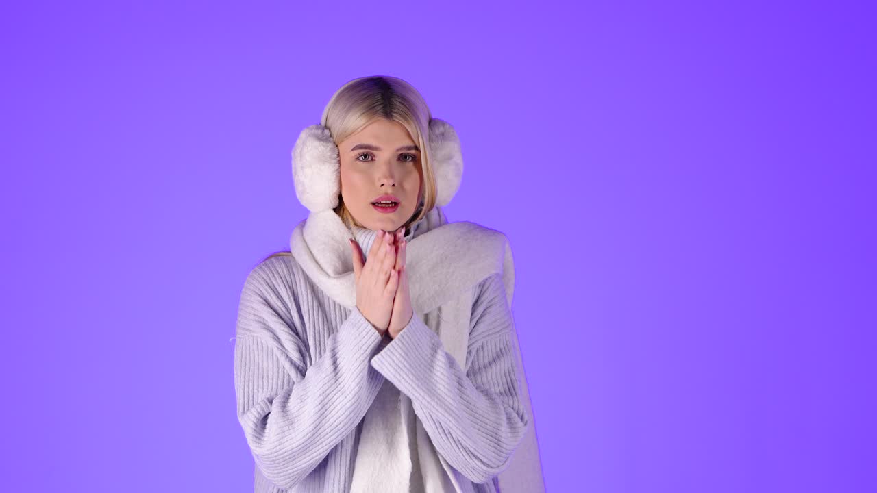 блондинка, чувствующая холод, потирает руки в рукава свитера, одета в зимнюю одежду, снимается в студии на фиолетовом фоне