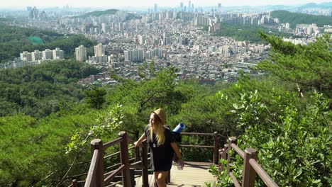 young caucasian girl climbing up on the trail while holding on the wooden railings with a city view from the gwanaksan mountain on a sunny day in seoul, south korea