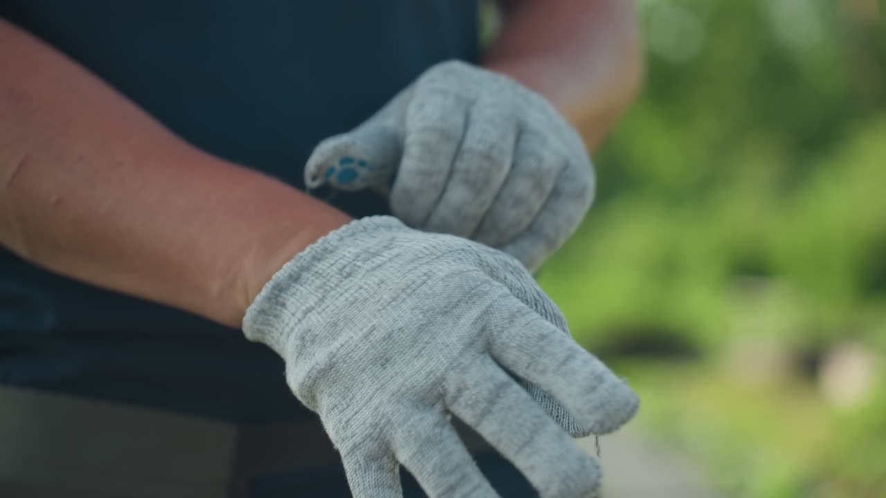 close up of farmer putting on ash color gloves, fingers slipping into fabric with precision, blurred green background, focus on texture and motion preparing for outdoor gardening task