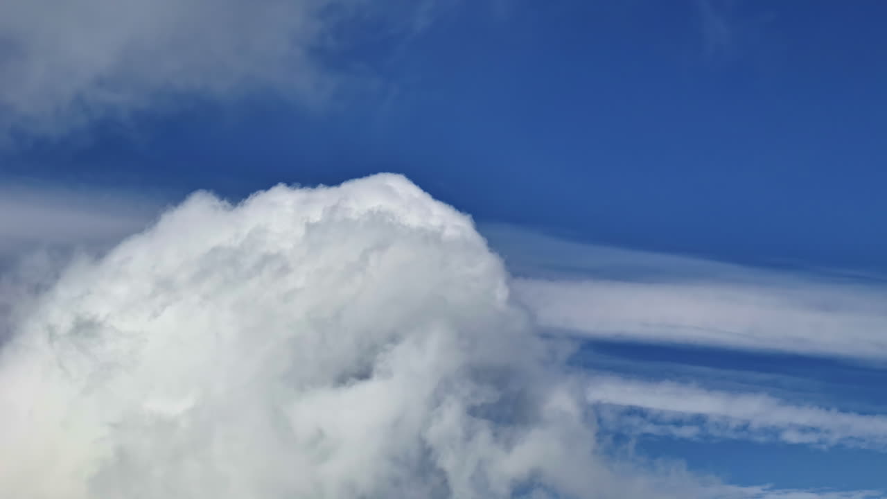 A beautiful and serene aerial shot from an airplane window, showing a flight past a huge, soft, white cumulus cloud formation against a vast, deep blue sky on a sunny day.