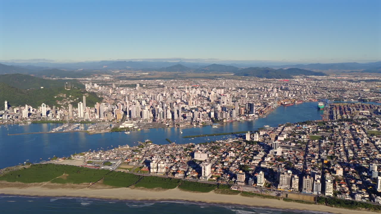 Backward drone retreats over Itajaí city, showing coastline, river, and mountains during afternoon. Sunny skies