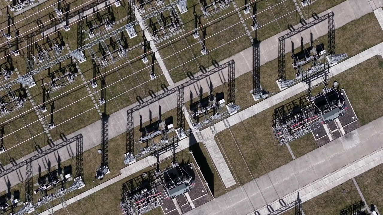 An aerial view shows an electrical substation facility with complex metal structures and transformers distributing power across a regional grid.