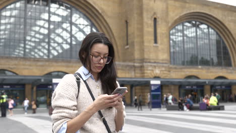 Attractive-young-woman-using-her-smartphone-while-stood-waiting-outside-the-main-entrance-to-Kings-Cross-Station,-London