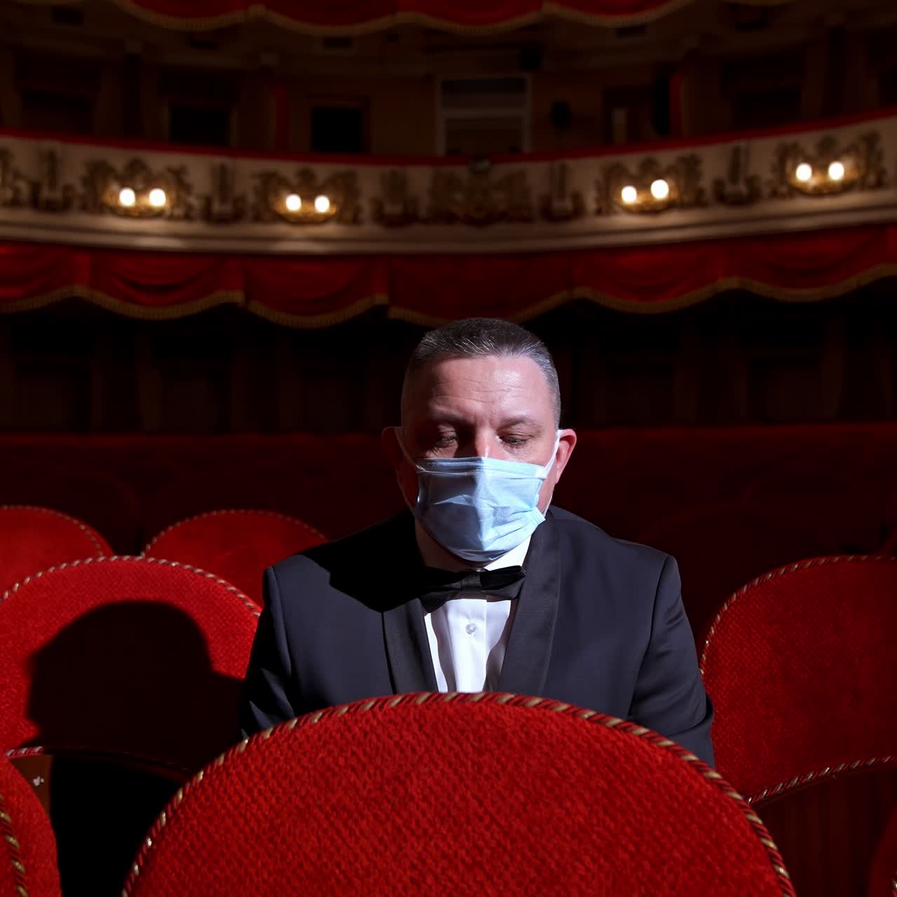 Visitor in suit and medical mask sitting in comfortable chairs alone on dark theater background. Male among rows of empty red chairs. Pandemic concept