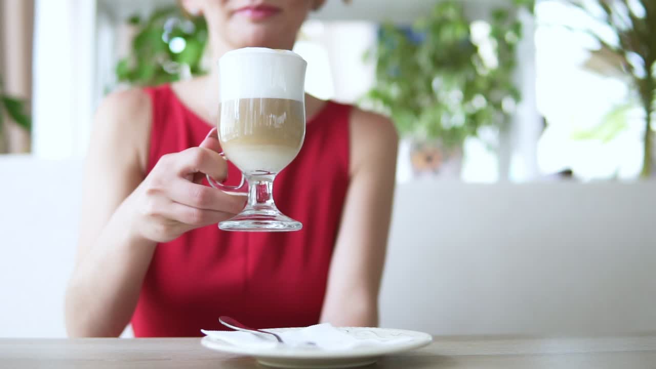 빨간색 조합의 긴 금발 머리카락을 가진 예쁜 젊은 여성이 카페에 앉아 늦게 마시고 누군가를 기다리고 있습니다. 느린 움직임