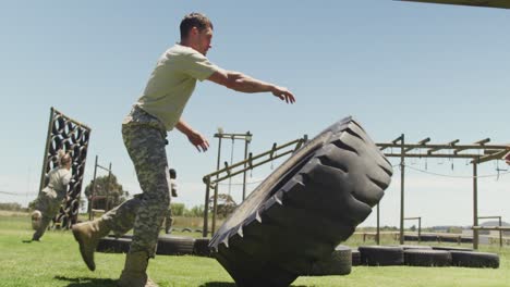 fit caucasian male soldier in army fatigues flipping tractor tyre on army obstacle course in the sun