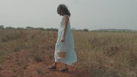 Slow-motion-handheld-shot-of-a-pretty-young-indian-woman-standing-on-a-dry-field-with-grass-dressed-in-light-blue-dress-and-carrying-a-wood-basket-during-windy-weather