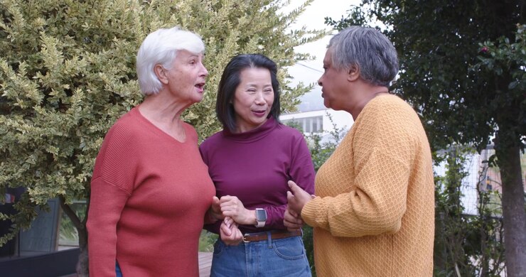 Diverse trio at porch, left senior woman speaking, center woman holding hands supporting smartwatch
