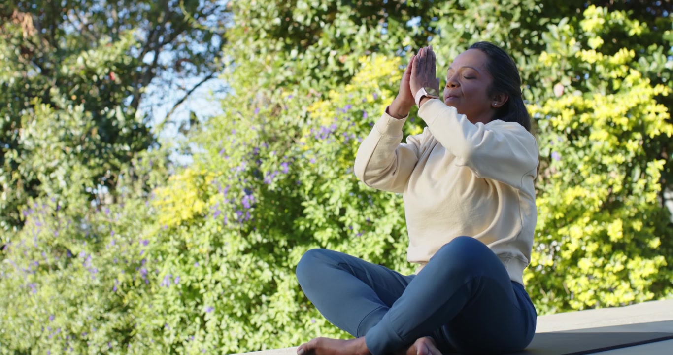 Mature African American woman sitting on mat ledge, inhaling, lifting arms, meditating, copy space