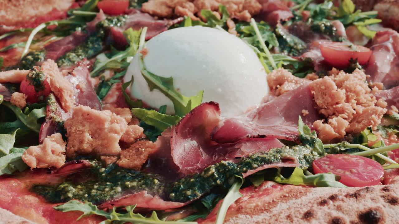 A beautifully shot overhead view of a gourmet pizza topped with burrata, prosciutto, cherry tomatoes, pesto, arugula, and crunchy crumbs