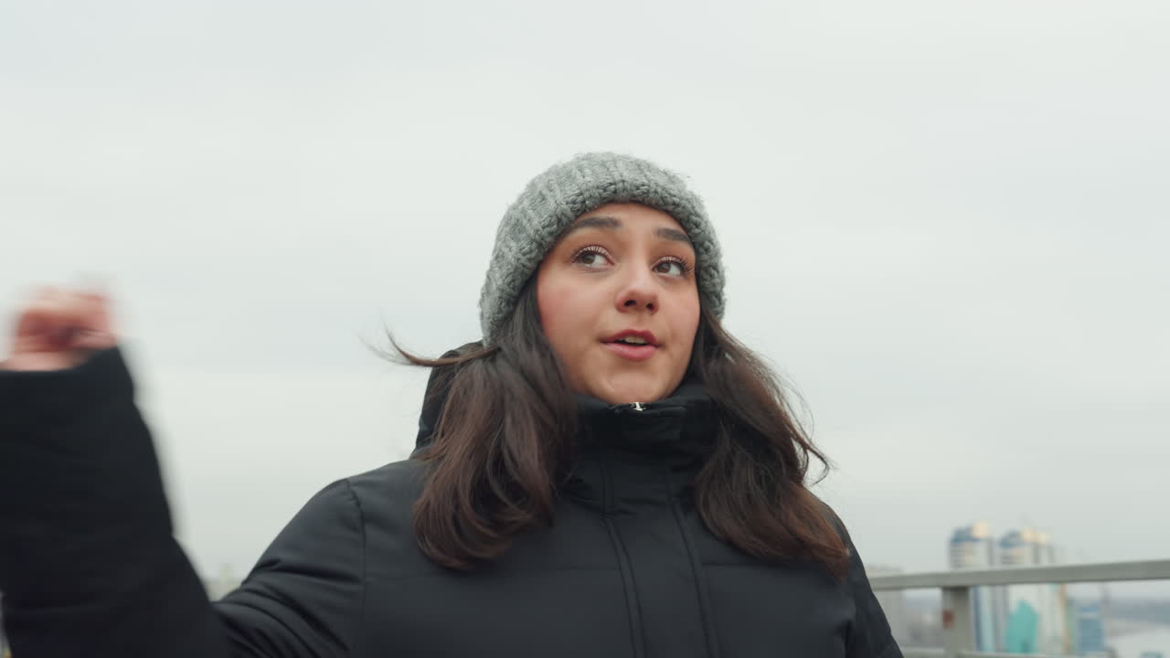 Bridge dance by river overlook, woman in knit hat practices measured arm movements and contemplative gaze across water, windtossed hair and railing foreground, muted palette and urban architecture