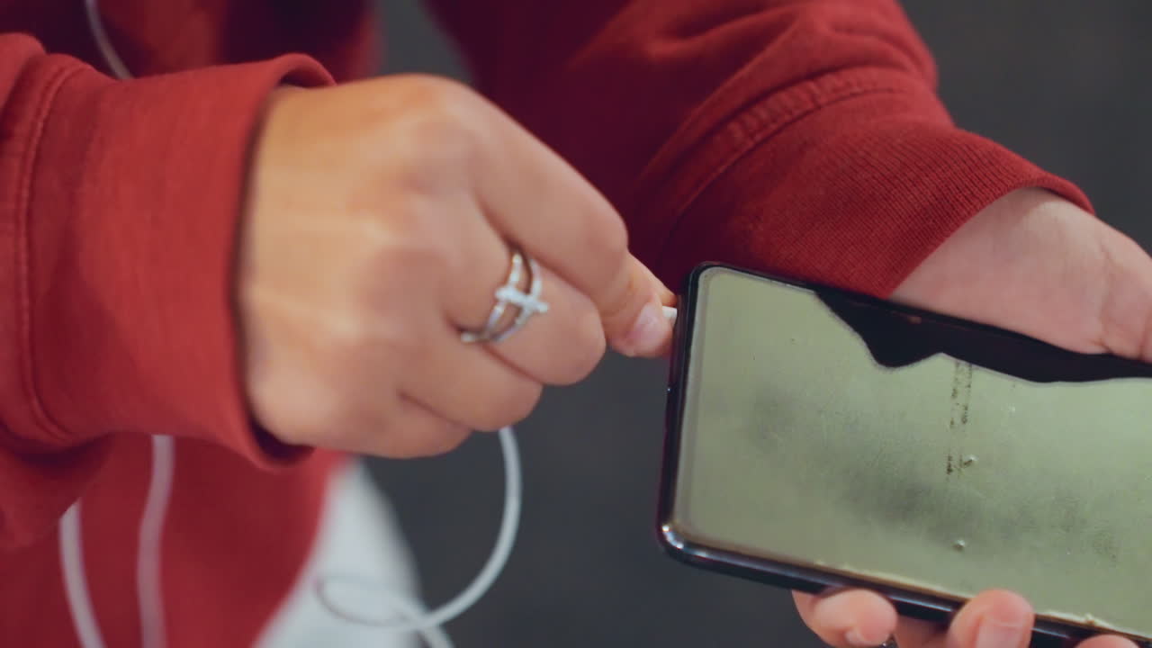 Closeup hands holding cracked smartphone, red hoodie, white earphone cable, plugging earbud into port, silver rings on fingers, urban evening commute, student connects playlist while walking, casual