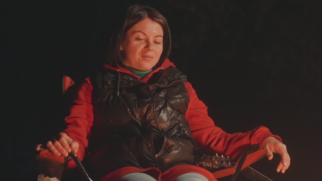 Quiet campfire scene with white woman warming hands, red hoodie under black puffer vest, seated in camp chair, ember glow on face, contemplative expression, night forest backdrop, cozy camping mood