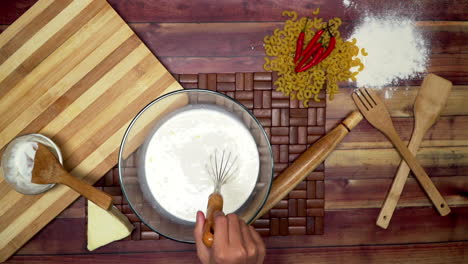 a top view of mixing the milk in the bowl, raw macaroni, wooden spoon, wooden spatula, flour, rolling pin and chili on the table