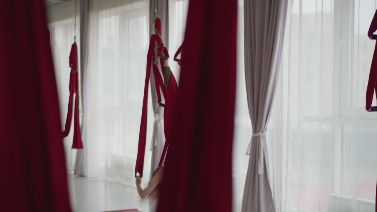 Fitness coach in sportswear performs inverted aerial silk pose in bright studio lined with red hammocks and sheer curtains, showing strength flexibility and serene wellness flow with swing