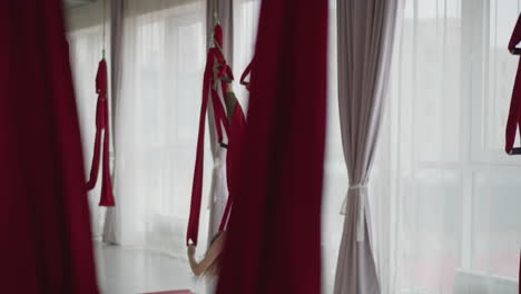 fitness coach in sportswear performs inverted aerial silk pose in bright studio lined with red hammocks and sheer curtains, showing strength flexibility and serene wellness flow with swing