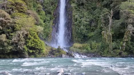 Großer-Wasserfall-Mit-Einem-Gletscherfluss-Davor,-Umgeben-Von-üppigem-Grünem-Regenwald-An-Der-Westküste-Neuseelands