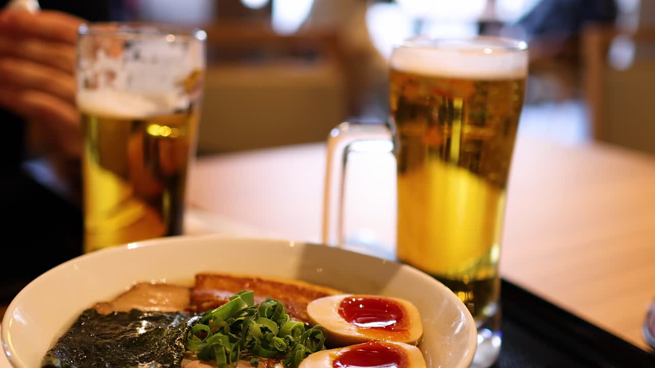 Close-up of a bowl of ramen with garnishes and a glass of beer on a wooden table.