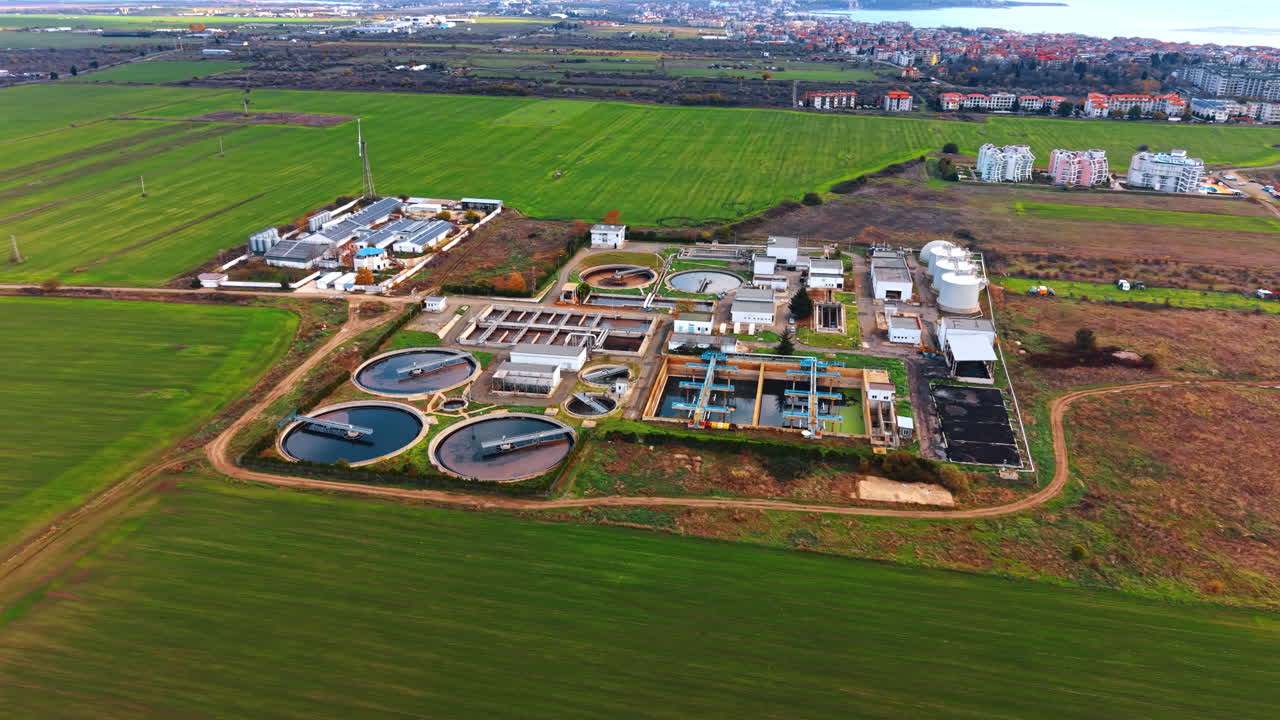 Vast green fields surround the territory of water treatment plant. Drone flight around the sewage recycling facility. Cityscape at backdrop