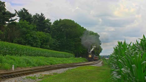 una vista frontal de un tren de pasajeros de vapor antiguo que se acerca viajando a través de tierras de cultivo y campos de maíz que soplan humo en un día soleado de verano