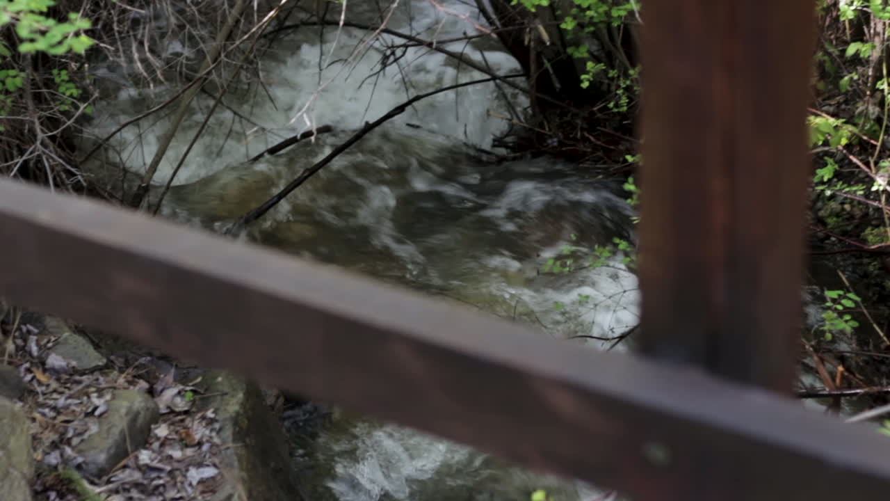 mirando más allá de la barandilla del puente de madera en una pequeña cascada de arroyo corriendo sobre rocas en el bosque de primavera